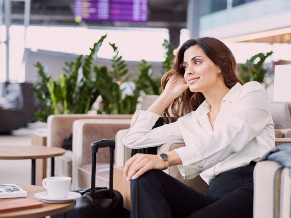 Eine Frau in Business-Kleidung sitzt in einem hellen Lounge-Sessel. Neben ihr steht ein Koffer, auf dem Tisch vor ihr befinden sich Reisedokumente und eine Tasse mit Untersetzer.