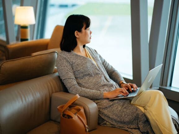 Eine Frau sitzt mit Laptop in einem Ledersessel am Fenster einer Flughafen-Lounge und schaut aufs Rollfeld.