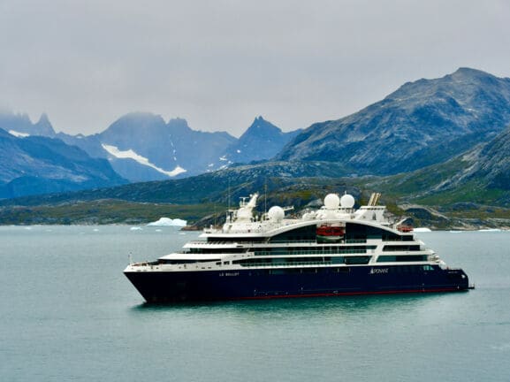 Das Bild zeigt im Vordergrund das Kreuzfahrtschiff. Im Hintergrund sind Berge und etwas Schnee und Eis zu sehen.