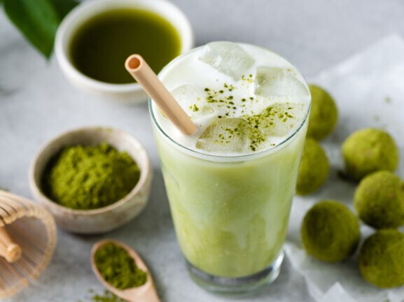 Ein Glas mit Matcha Latte und Strohhalm, daneben liegen grüne Kugeln, eine Schale mit grünem Pulver, ein Bambusbesen und eine Schale mit purem Matchatee.