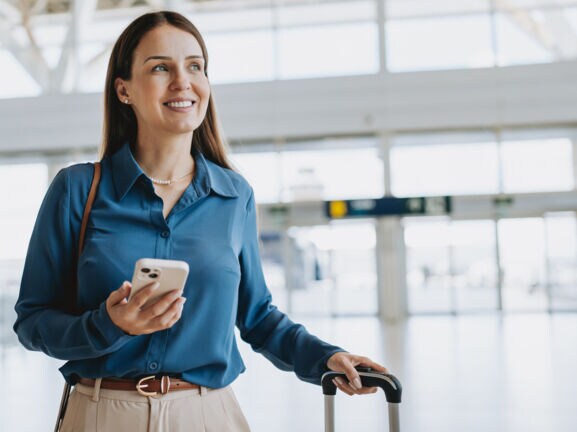 Eine Frau am Flughafen, die in einer Hand einen Koffergriff hält und in der anderen ein Smartphone.