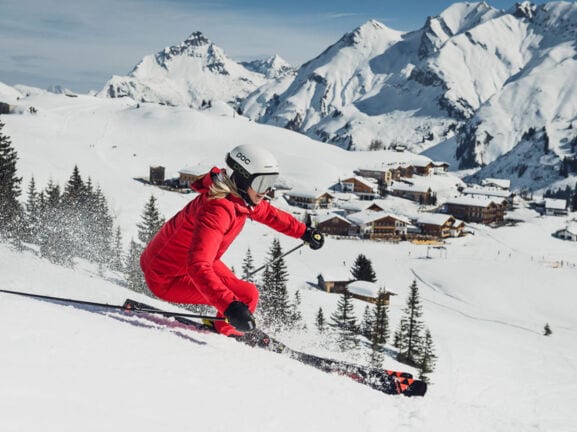 Eine Skifahrerin im roten Schneeanzug bei der Abfahrt, im Hintergrund schneebedeckte Chalets vor Bergpanorama.