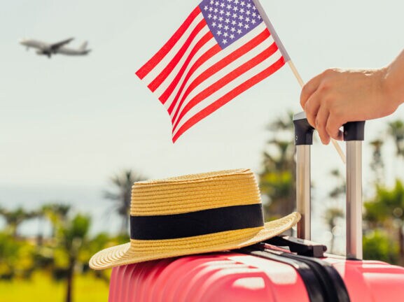 Ein Strohhut auf einem pinken Hartschalenkoffer, eine Hand am Griff hält eine kleine US-amerikanische Flagge, im Hintergrund Plamen und ein Flugzeug am Himmel.