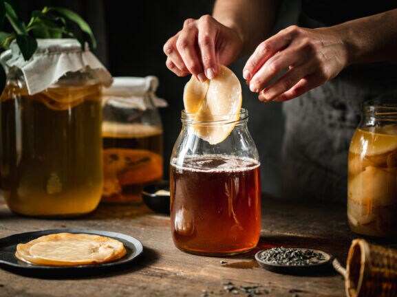 Ein Kombucha-Pilz wird von einer Hand in ein Gefäß mit brauner Flüssigkeit auf einem Holztisch gegeben, daneben weitere befüllte Einmachgläser.