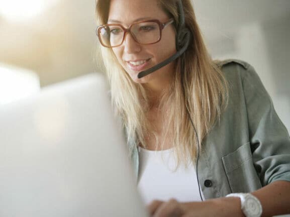 Eine blonde Frau mit Headset sitzt vor einem Laptop und arbeitet.