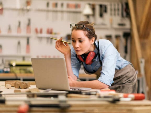 Foto einer Handwerkerin, wie sie an ihrer Werkbank steht und auf den vor ihr geöffneten Laptop blickt.