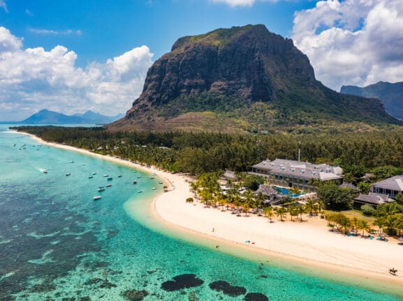 Küstenpanorama mit Hotelresort an einem palmengesäumten, weißen Sandstrand an türkisblauem Meer, im Hintergrund Berge.