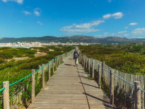 Eine unkenntliche Person beim Wandern in Portugal auf einem Holzsteg, der in Küstennähe über grüne Vegetation führt.