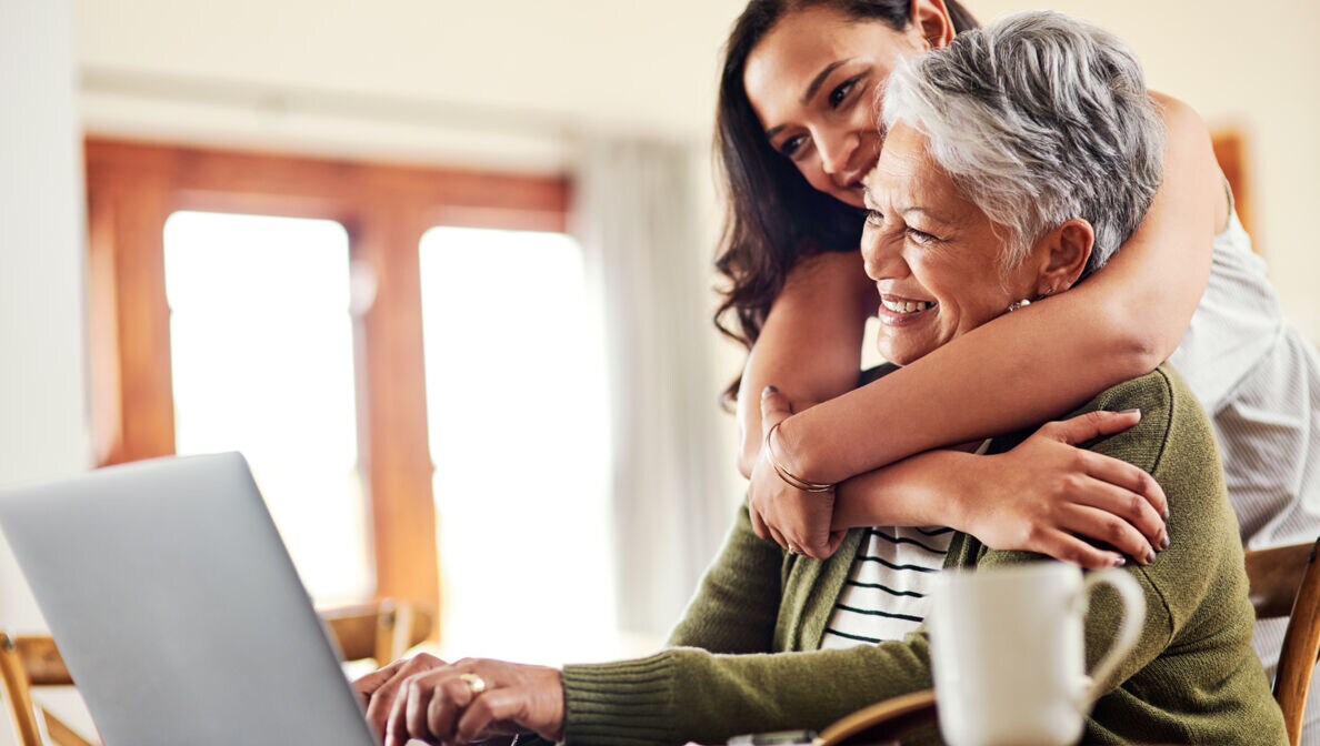 Eine ältere Frau sitzt am Tisch vor einem Laptop und lächelt. Eine jüngere Frau umarmt sie.