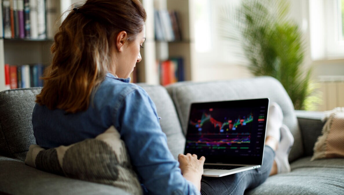 Eine junge Frau sitzt mit Laptop auf ihrem Sofa und prüft Aktienkurse