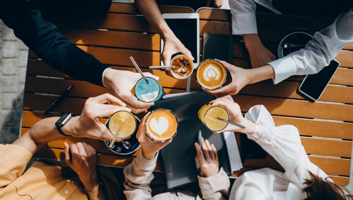 Table-top-Ansicht einer Gruppe von jungen Menschen, die mit verschiedenen Getränken in der Hand anstoßen. Auf dem Tisch darunter liegt ein Tablet, ein Smartphone und Dokumente.
