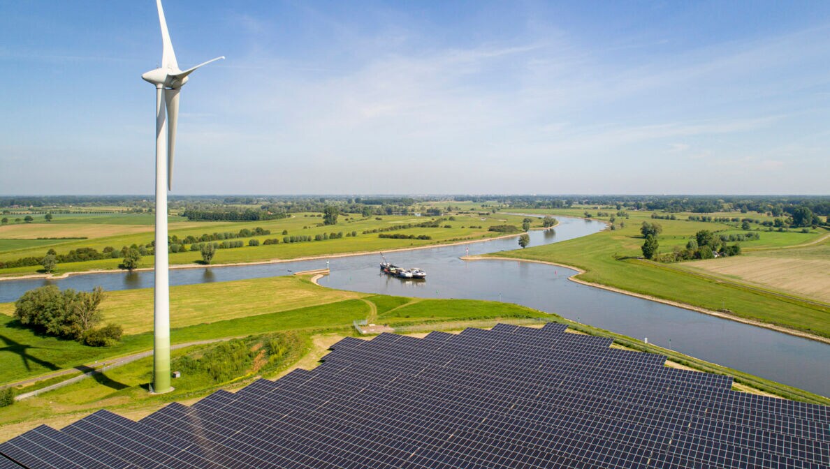 Blick auf eine grüne Landschaft und einen Fluss hinter einem Windrad und einer großen Solaranlage