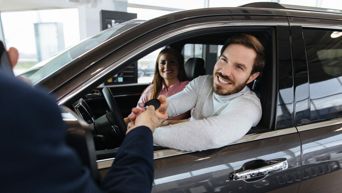 Eine Frau in einem Auto neben einem Mann, der von einer Person durch das Autofenster einen Autoschlüssel annimmt