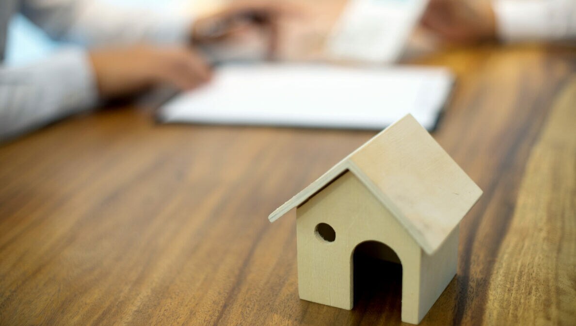 Ein kleines Hausmodell aus Holz steht auf einem Schreibtisch, im Hintergrund sind unscharf zwei Personen und Unterlagen zu erkennen.