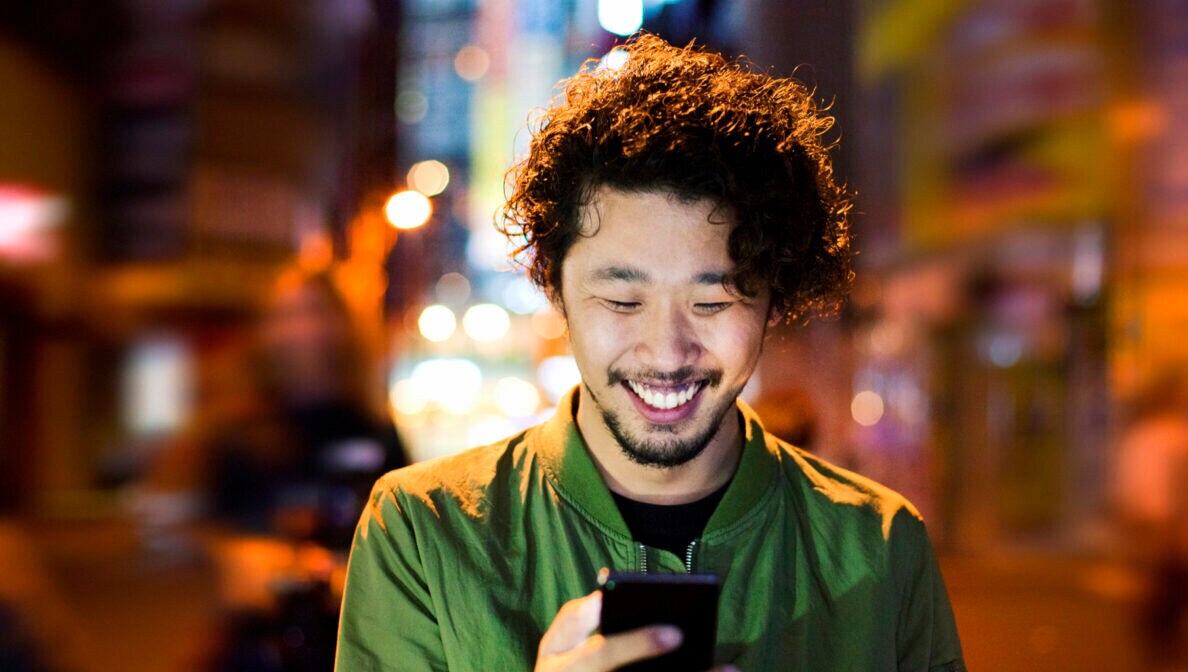 Ein Mann mit Smartphone in der Hand, in dessen Richtung er lächelt. Im Hintergrund eine verschwommene Großstadtkulisse.