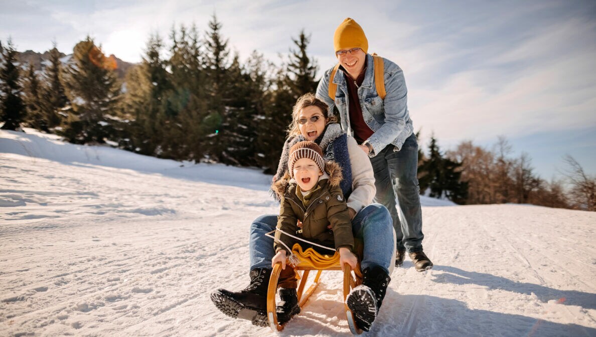 Eine dreiköpfige Familie im Schnee, von der zwei Personen auf einem Schlitten sitzen und die dritte Person anschiebt.