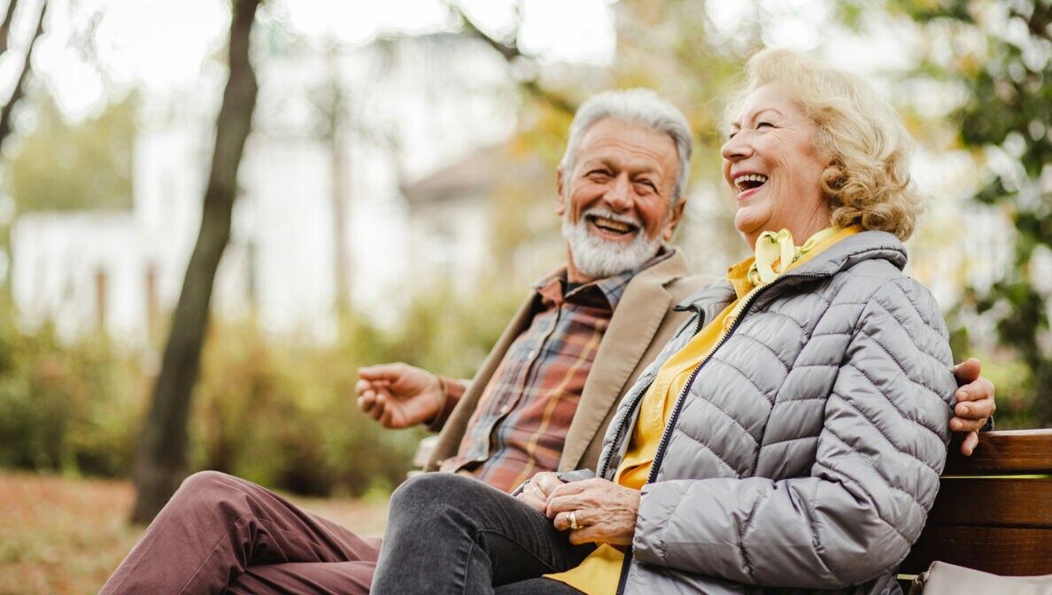 Zwei ältere Menschen sitzen lachend auf einer Parkbank.