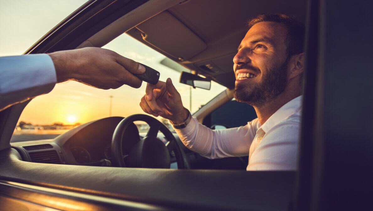 Ein lächelnder junger Mann auf dem Fahrersitz im Auto. Eine weitere Person reicht ihm durch das geöffnete Autofenster einen Autoschlüssel
