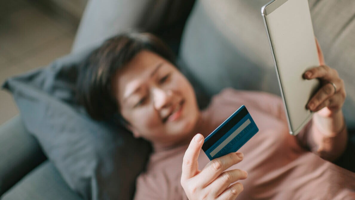 Eine Person liegt auf der Couch. Sie hält in der einen Hand eine Kreditkarte und in der anderen ein Smartphone.