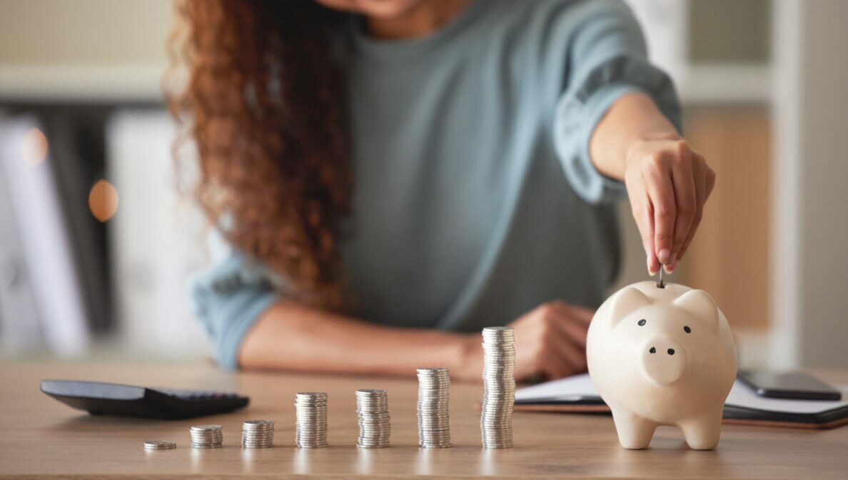 Eine junge Frau steckt eine Geldmünze in ein weißes Sparschwein, das auf einem Tisch steht. Daneben befinden sich verschieden große Münzstapel und ein Taschenrechner.