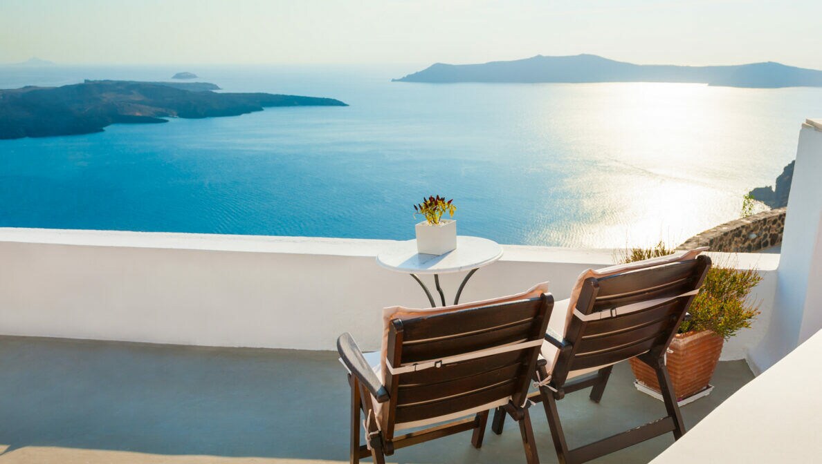 Zwei Holzstühle stehen auf einer sonnigen Terrasse mit Blick übers Meer.