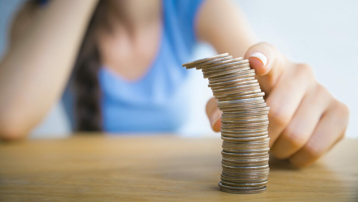 Eine Person sitzt am Tisch. Auf dem Tisch sind Münzen aufgestapelt. Mit einem Finger bringt die Person den Stapel ins Kippen.
