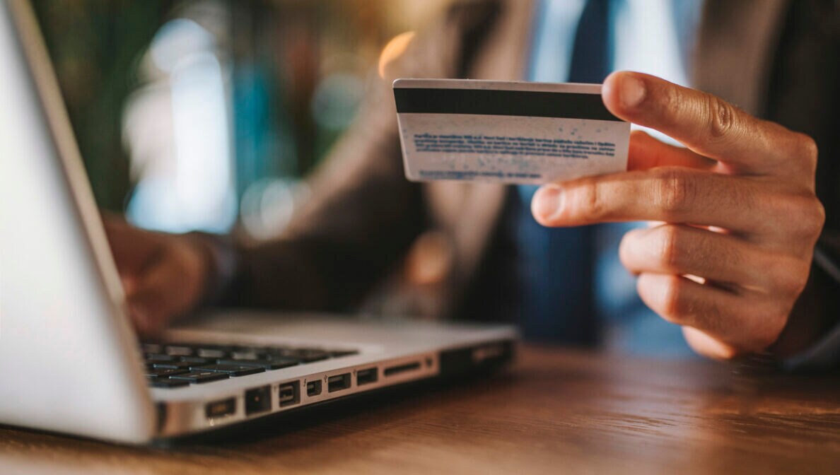 Nahaufnahme einer Hand, die neben einem geöffneten Laptop eine Kreditkarte in der Hand hält.