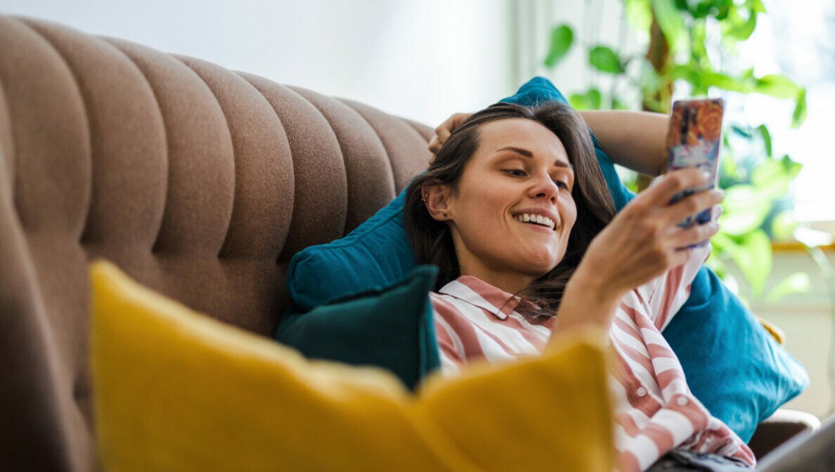 Eine Person liegt auf dem Sofa und schaut lächelnd auf ihr Smartphone.