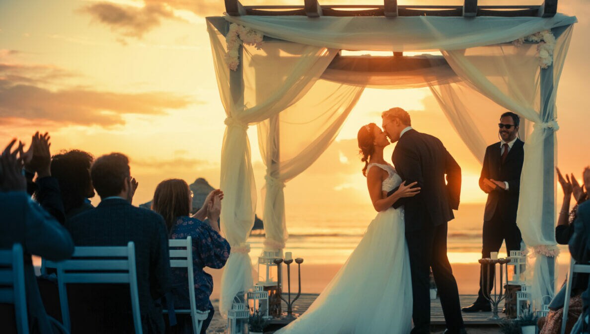 Ein Brautpaar steht bei Sonnenuntergang unter einem Baldachin im Freien und küsst sich. Mehrere Gäste sitzen auf Stühlen und klatschen
