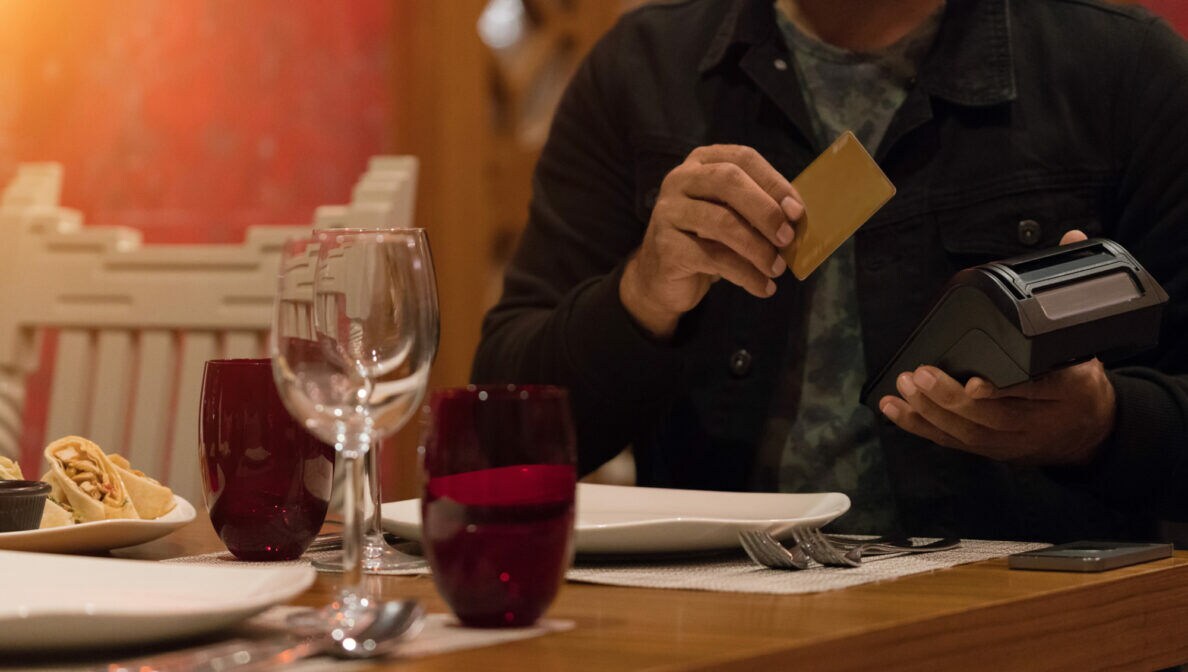 Nahaufnahme einer Person an einem Tisch im Restaurant. Sie hält ein Kartenlesegerät in der einen und eine Kreditkarte in der anderen Hand.
