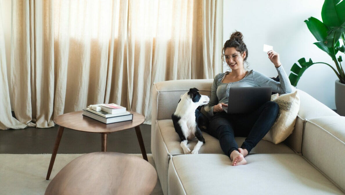 Eine Frau sitzt mit ausgestreckten Beinen auf einem Sofa, neben ihr sitzt ein Hund. Auf ihrem Schoß ist ein Laptop und in einer Hand hält sie eine Kreditkarte