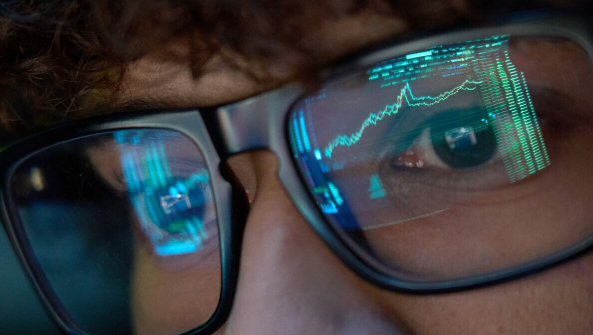 Eine Person schaut auf einen Monitor. In ihrer Brille spiegeln sich die Charts von Aktienkursen.