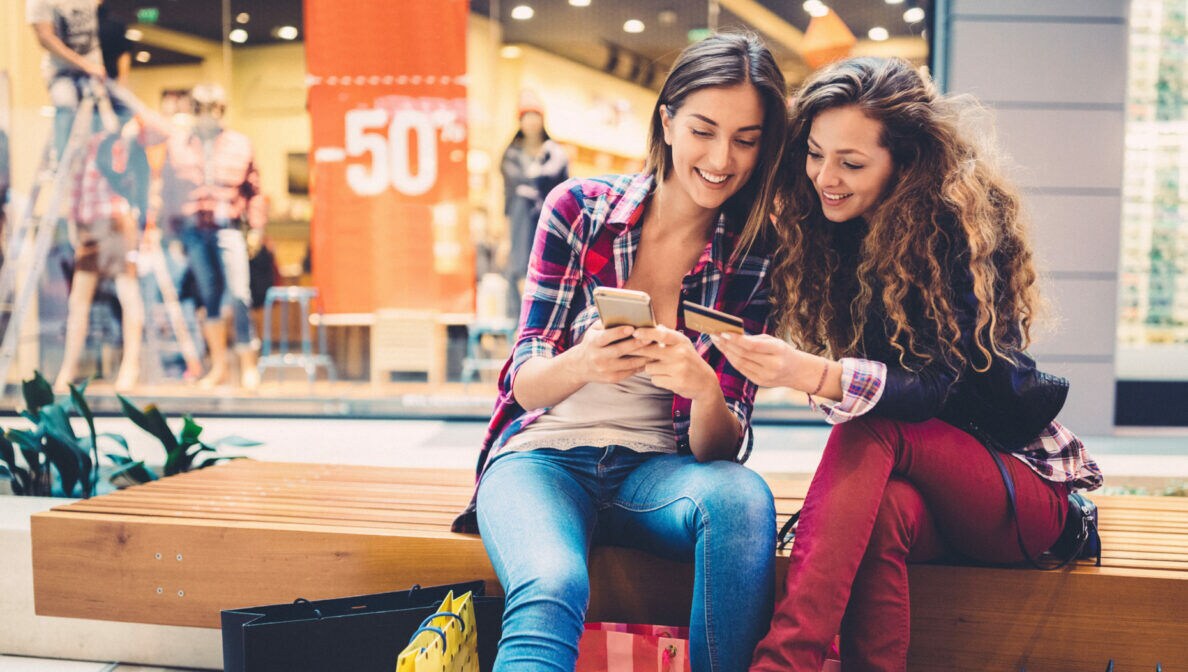 Zwei Personen sitzen in einer Shoppingmall auf einer Bank und schauen gemeinsam auf ein Smartphone und eine Kreditkarte.