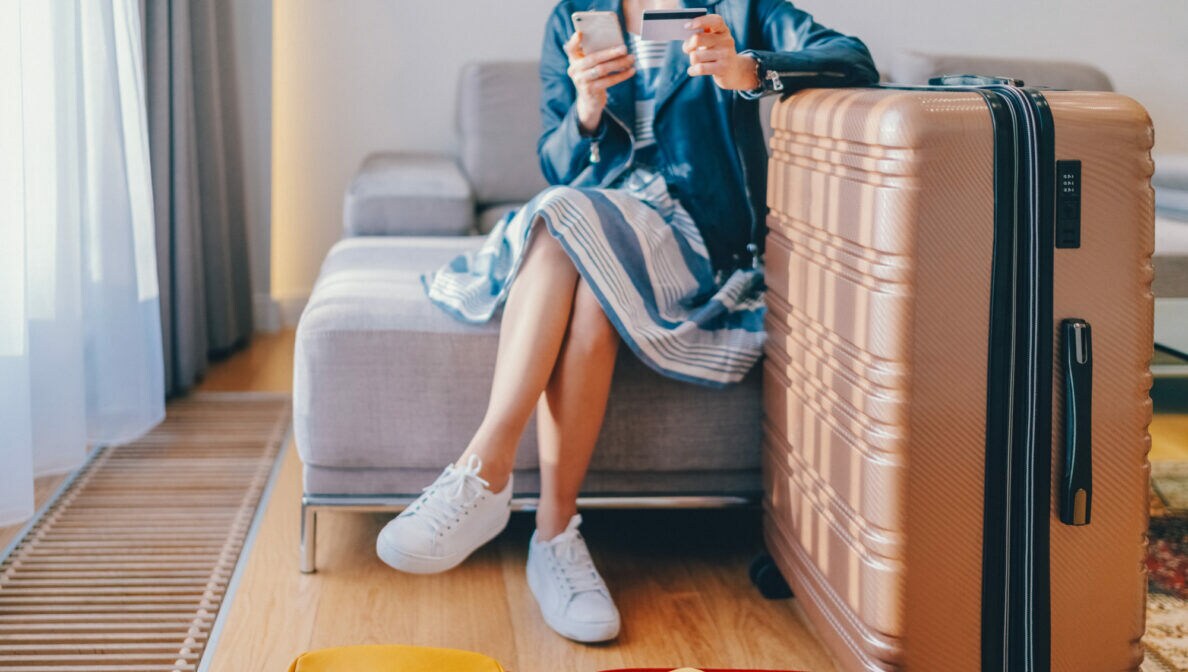 Eine Person, dessen Kopf nicht zu sehen ist, sitzt auf einem Sofa, vor ihr befinden sich zwei Koffer und sie hält ein Smartphone und eine Bankkarte in den Händen.