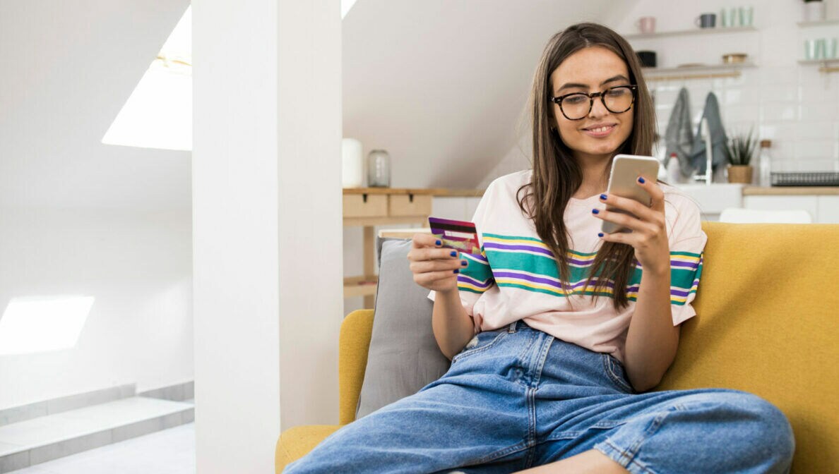 Eine Jugendliche mit langen braunen Haaren und Brille sitzt auf einer Couch und schaut lächelnd auf ein Smartphone in ihrer rechten Hand. In der linken Hand hält sie eine Bankkarte.