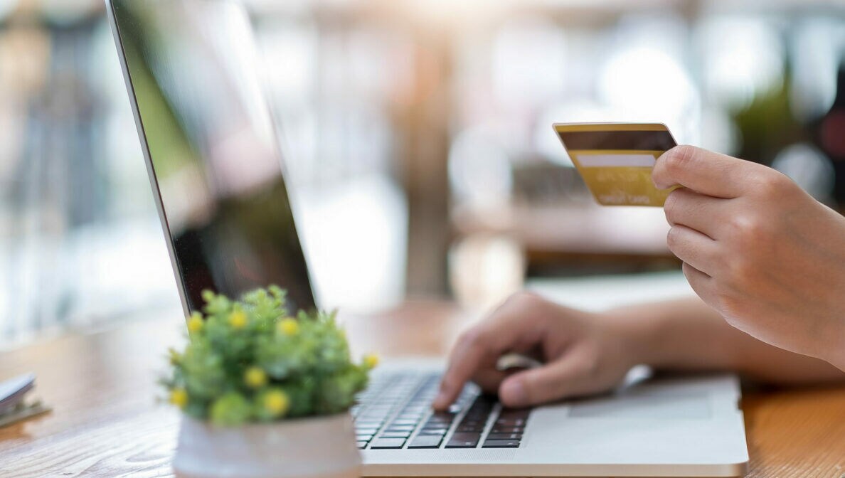 Nahaufnahme einer Hand, die eine Bankkarte hält, die andere Hand liegt auf einer Laptoptastatur.