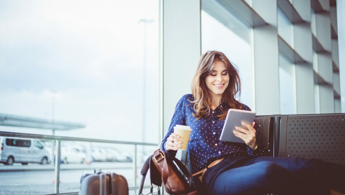 Eine Person sitzt am Flughafen und hält einen Kaffeebecher und ein Tablet in den Händen. Ein Koffer steht neben ihr