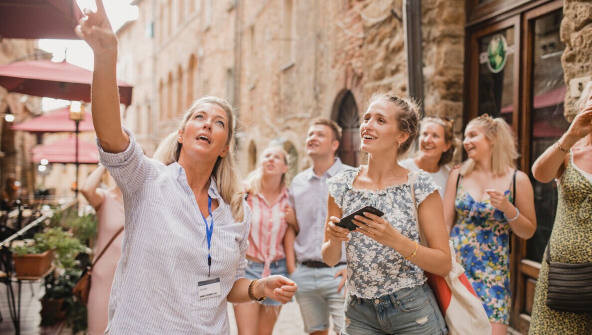 Eine Gruppe von Menschen steht in der Straße einer Altstadt. Sie hören einer Reiseleiterin zu.