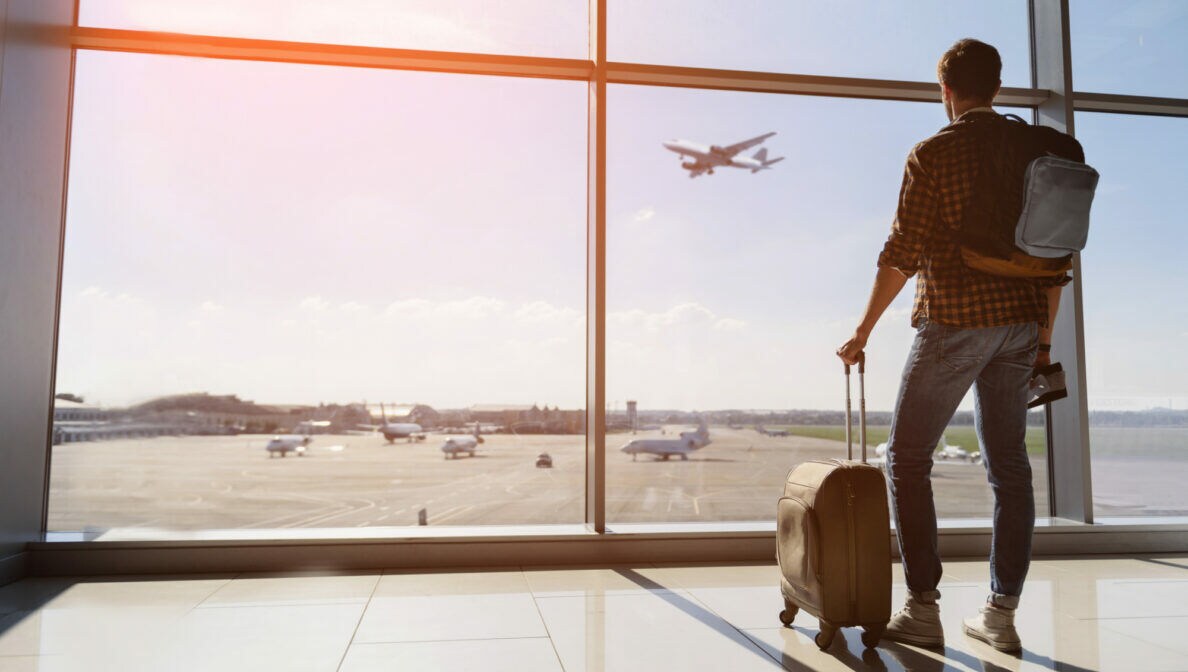 Ein Mann mit Rucksack und Koffer steht am Glasfenster, hinter dem Flugzeuge zu sehen sind