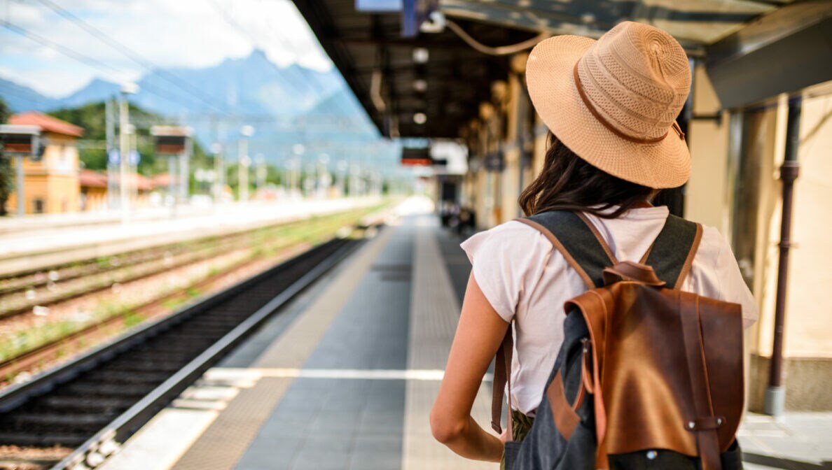 Eine Person in Rückansicht mit Rucksack und Hut steht am Bahnsteig.