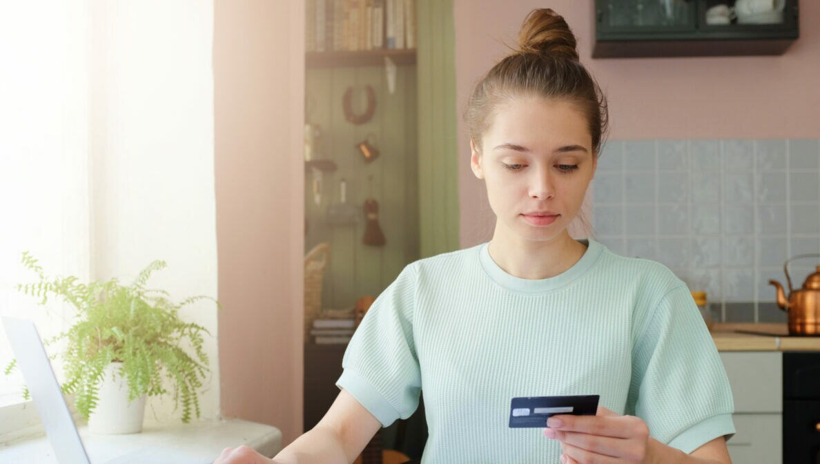 Eine Person sitzt am Küchentisch, schaut auf ihre Bankkarte und hat mehrere Dokumente vor sich liegen sowie die andere Hand auf einem geöffneten Laptop.