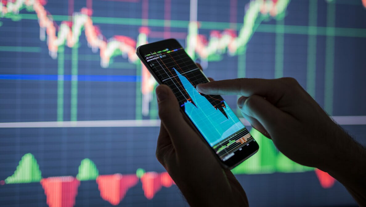 Nahaufnahme von zwei Händen, die ein Smartphone mit Aktienkurs halten. Im Hintergrund ist ein großes Display mit weiteren Kursen zu erkennen.