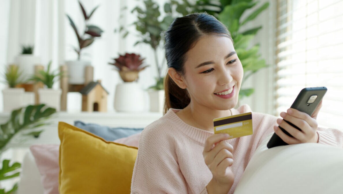 Eine Person sitzt auf einem Sofa, halt eine Bankkarte in der einen Hand und blickt auf ihr Smartphone in der anderen Hand.