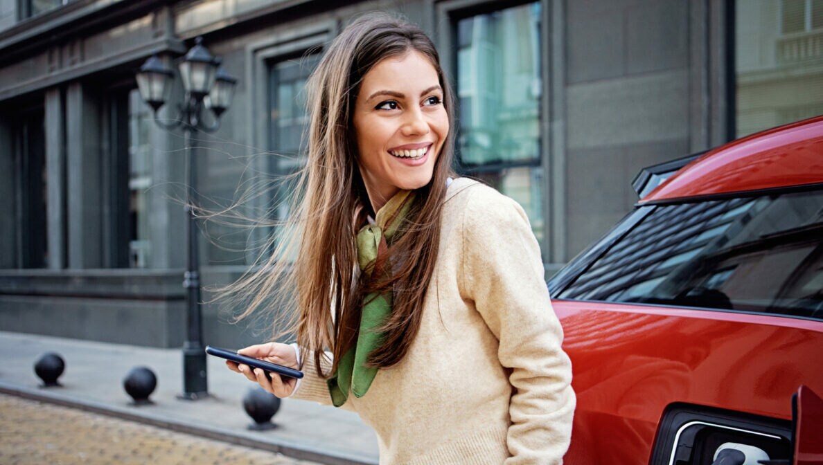 Eine Person steht vor einem roten E-Fahrzeug und hält ein Smartphone in der Hand.