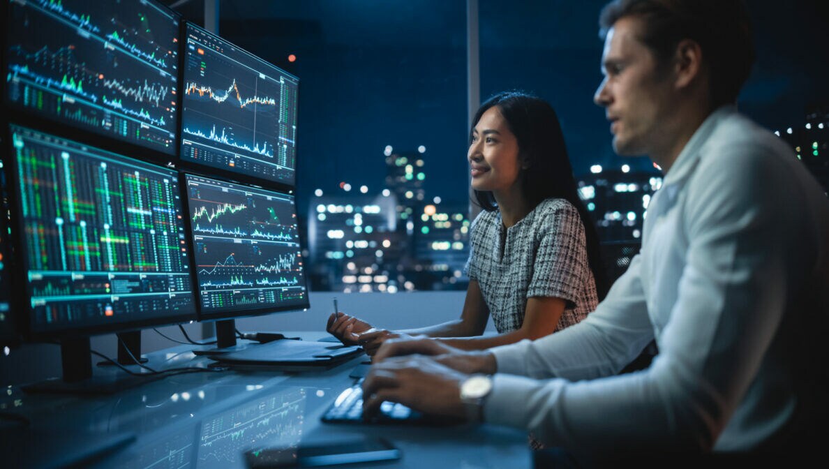 Zwei Personen sitzen in einem Büro in einer Großstadt am Schreibtisch, auf den vier Bildschirmen sind Börsencharts, Indikatoren und weitere Daten zu sehen.