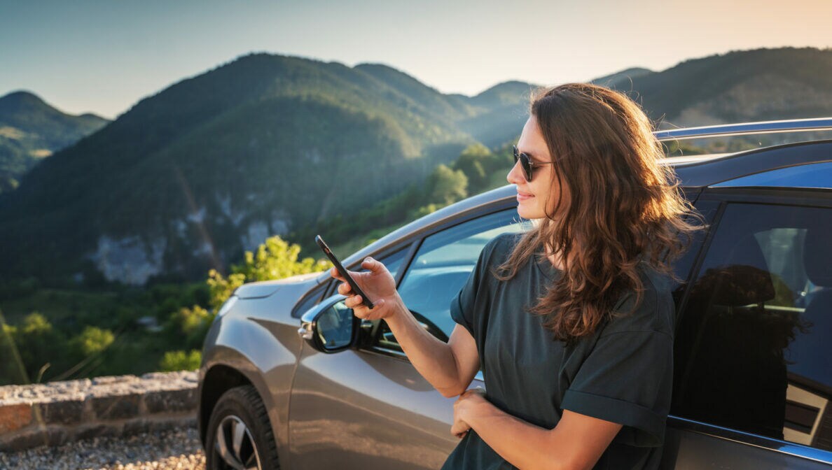 Eine Person lehnt an einem Auto und blickt auf ein Smartphone, im Hintergrund sind Berge zu erkennen.