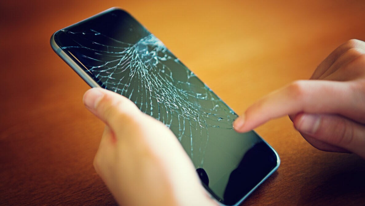 Eine Person hält ein Handy mit zersprungenem Display in der einen Hand und tippt mit einem Finger der anderen Hand darauf.