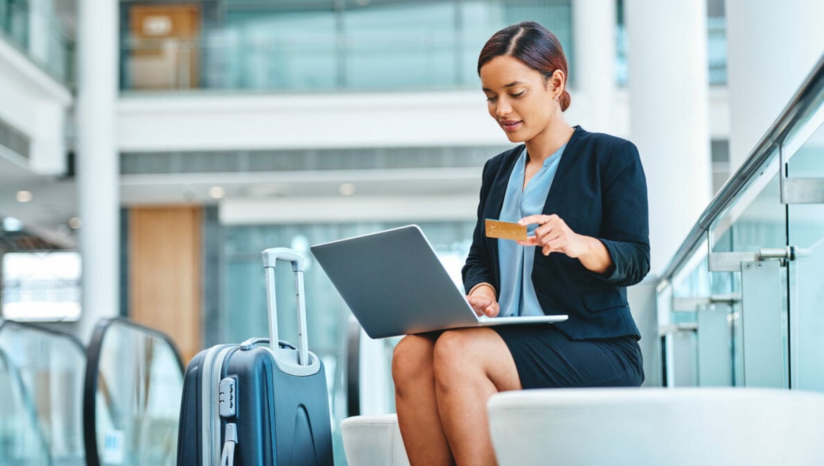 Eine Person sitzt in einem Bürogebäude, hat einen Koffer neben sich stehen und ein Laptop auf den Knien. In ihrer Hand hält sie eine Bankkarte.