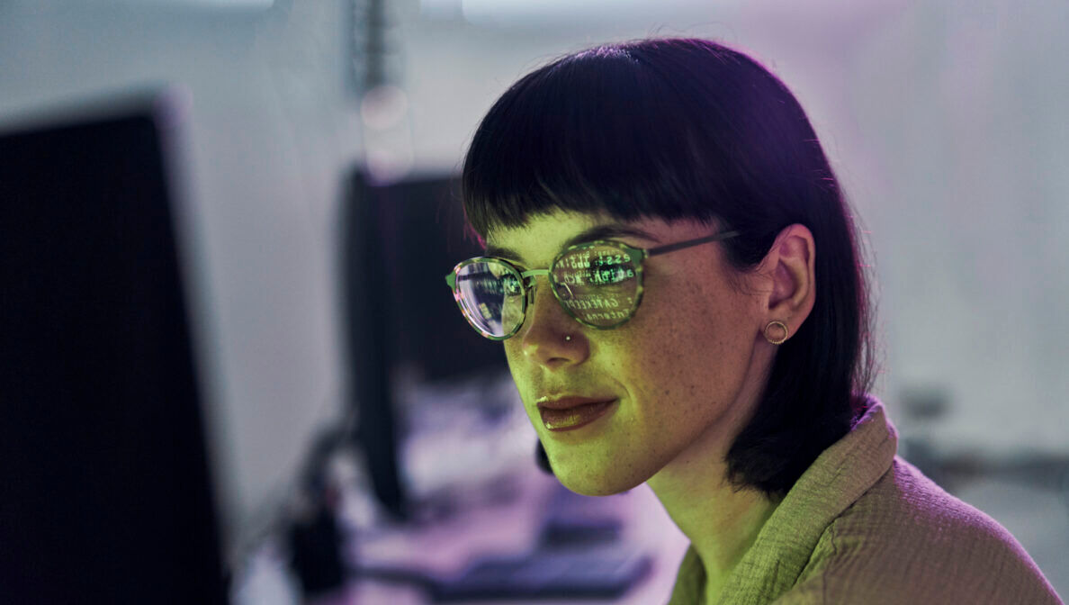 Portraitaufnahme einer Frau mit Brille, in deren Gläsern sich die Schrift eines Computerbildschirms widerspiegelt.