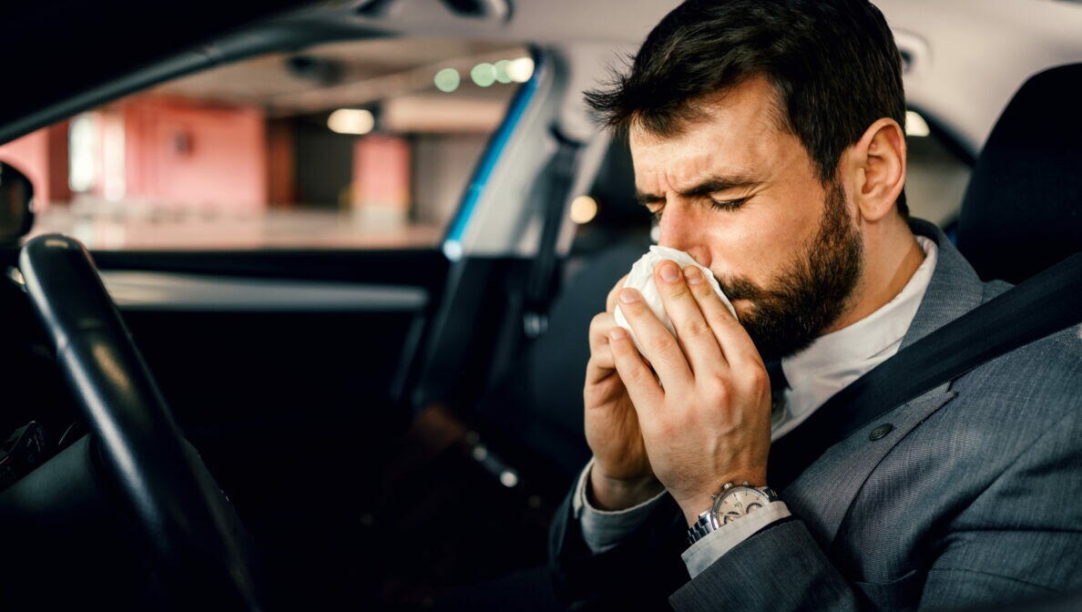 Ein dunkelhaariger Mann im Anzug sitzt angeschnallt am Steuer eines parkenden Autos und putzt seine Nase.
